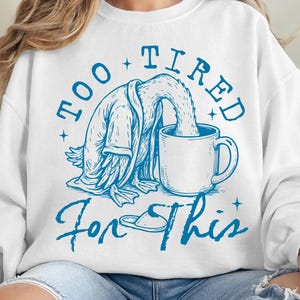 Op de afbeelding: Witte trui met een blauwe afbeelding van een eend die een badjas draagt en in een kop koffie valt. De tekst "Too Tired For This" is in het blauw geschreven.
