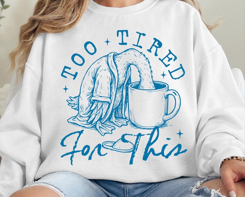 Op de afbeelding: Witte trui met een blauwe afbeelding van een eend die een badjas draagt en in een kop koffie valt. De tekst "Too Tired For This" is in het blauw geschreven.