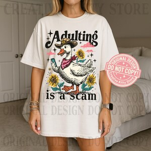 May include: A cream-coloured t-shirt with a cartoon goose wearing a cowboy hat and bandana, holding a phone. The text "Adulting is a scam" is below the goose, with sunflowers and pink clouds in the design.
