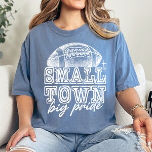 May include: A blue t-shirt with a white vintage-style graphic of a football and the text "SMALL TOWN big pride".
