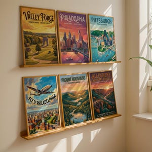 May include: Six framed travel posters with wooden frames are displayed on a wall. The posters feature vintage-style illustrations of Pennsylvania locations, including Valley Forge, Philadelphia, and Pittsburgh. The bottom row includes a poster that says "Fly to Philadelphia".