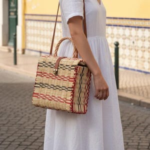 Può includere: Borsa a mano rettangolare in paglia intrecciata con manico superiore. La borsa presenta strisce rosse, nere e color naturale. Ha un cinturino in pelle marrone e una chiusura con fibbia. Lo sfondo mostra un edificio giallo e una strada acciottolata.