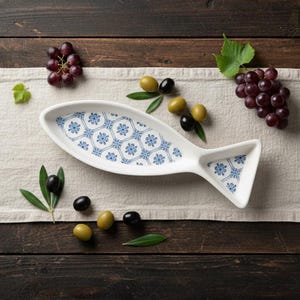 May include: A white ceramic fish-shaped serving dish with a blue floral pattern, displayed on a linen cloth. Grapes and olives are arranged around the dish on a dark wooden surface, creating a rustic, Mediterranean-inspired aesthetic.