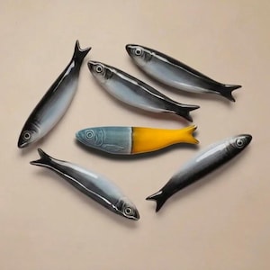 May include: Six ceramic fish figurines. Five are gray, white, and black, with a realistic design. One fish is half blue and half yellow. The fish are arranged on a beige surface.