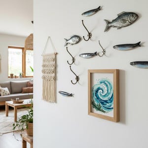 May include: A coastal-themed wall arrangement featuring metal fish sculptures, decorative hooks, and a framed watercolor print of a wave. A macrame wall hanging adds texture. The decor is set against a white wall, creating a serene, nautical ambiance.