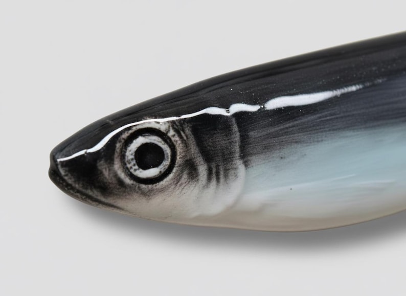 Pu&ograve; includere: Primo piano di una testa di pesce in ceramica. Il pesce &egrave; dipinto in tonalit&agrave; di nero, bianco e blu. L'occhio &egrave; un cerchio scuro con un contorno bianco. Il corpo del pesce &egrave; liscio e curvo, con una striscia bianca sulla parte superiore.
