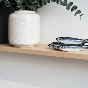 May include: A decorative shelf arrangement featuring three ceramic fish figurines with blue and white details. A white crackle-glazed vase and antique books are also on the shelf, with greenery adding a natural touch.