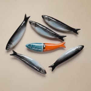 May include: A collection of ceramic fish figurines. The fish are primarily black, white, and blue, with one fish featuring a blue and orange design. The figurines are arranged on a neutral-toned surface.