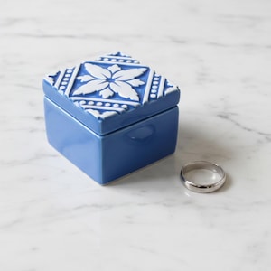 Può includere: Un portagioie in ceramica blu con un motivo floreale bianco sul coperchio, accanto a un anello d'argento. La scatola è quadrata con una finitura lucida. L'anello è una semplice fascia.