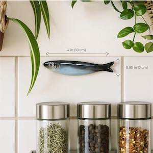 May include: A ceramic fish-shaped wall decor, measuring 4 inches (10 cm) long and 0.80 inches (2 cm) high, is mounted on a white tiled wall. The fish has a blue and gray color scheme. Below are three glass spice jars.