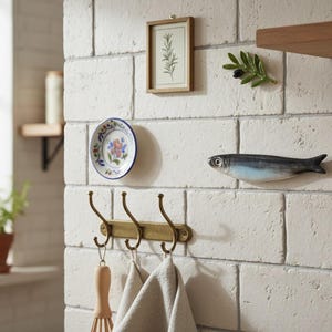 May include: A kitchen wall scene featuring decorative elements. A small framed botanical print, a ceramic plate with floral design, and a fish-shaped ornament are mounted on a white brick wall. A brass hook rack holds kitchen towels and a whisk.