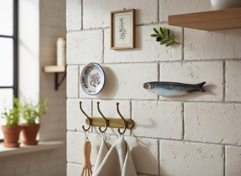 May include: A kitchen wall scene featuring decorative elements. A small framed botanical print, a ceramic plate with floral design, and a fish-shaped ornament are mounted on a white brick wall. A brass hook rack holds kitchen towels and a whisk.