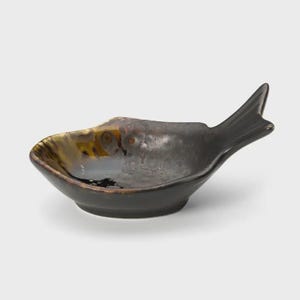 Könnte beinhalten: Eine kleine Keramikschale in Fischform mit glänzender Oberfläche. Die Schale hat eine dunkelbraune und schwarze Glasur mit gesprenkelten, metallisch wirkenden Highlights und eine glatte, geschwungene Innenfläche, die zum Schwanz zuläuft.