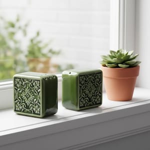 Puede incluir: Dos saleros y pimenteros de cerámica verde con diseños florales en relieve. Los saleros son de forma cuadrada y están sobre un alféizar blanco junto a una pequeña maceta de terracota con una planta suculenta.