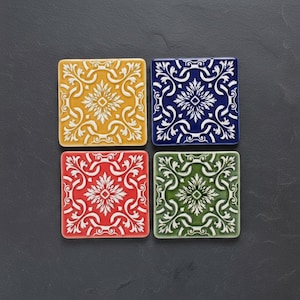 Könnte beinhalten: Vier quadratische Keramikuntersetzer mit erhabenem floralem Design. Die Untersetzer sind in Gelb, Blau, Rot und Grün gehalten, jedes mit einem weißen Muster. Die Untersetzer sind etwa 10 cm groß.
