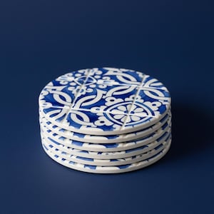 Può includere: Una pila di sei sottobicchieri rotondi in ceramica. Ogni sottobicchiere presenta un motivo floreale bianco e blu. I sottobicchieri sono impilati uno sopra l'altro, con il sottobicchiere superiore che mostra il design completo. Lo sfondo è blu scuro.