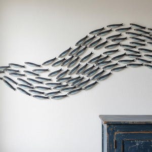 May include: A wall-mounted art piece featuring a school of fish. The fish are painted in shades of blue and gray, arranged in a flowing, wave-like pattern against a white wall. A dark blue cabinet is visible below.