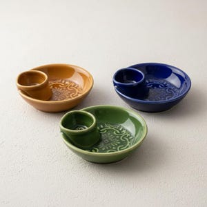 May include: Three ceramic serving dishes in mustard yellow, blue, and green. Each dish has a small, attached cup and a floral pattern. The dishes are designed for serving dips or sauces.