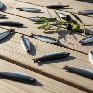 May include: Ceramic sardine figurines in various positions on a wooden surface. Some sardines are dark gray, others are light gray with white bellies. Olive branch with green and black olives adds a natural touch.