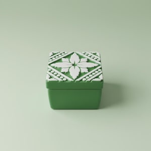 May include: A square, green decorative box with a white floral design on the lid. The lid features a raised, symmetrical pattern of a flower and geometric shapes. The box is set against a light green background.