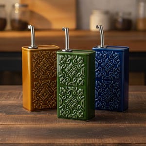 Puede incluir: Tres dispensadores de aceite de cerámica en amarillo mostaza, verde y azul marino. Cada botella rectangular presenta un patrón floral en relieve decorativo y una boquilla de acero inoxidable. Los dispensadores se muestran sobre una superficie de madera.