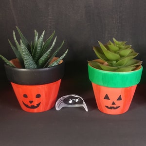 May include: Three small terracotta pots painted with jack-o'-lantern faces. The pots are filled with green succulents and have black rims. Two of the pots have black rims, and one has a green rim.
