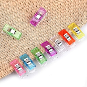 May include: A selection of multi-coloured sewing clips, including pink, blue, green, purple, orange, and yellow. Each clip has a clear body with a metal spring. They are arranged on a textured, tan fabric.