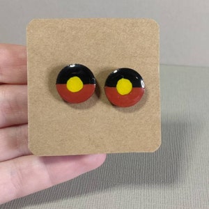 Op de afbeelding: Een paar kleine, ronde oorbellen met een zwart, geel en rood ontwerp. Het ontwerp lijkt op de Australische Aboriginalvlag.