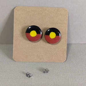 Op de afbeelding: Een paar oorbellen met een zwart, rood en geel Aboriginal vlagontwerp. De oorbellen liggen op een bruine kaart met twee zilveren oorbelsluitingen.