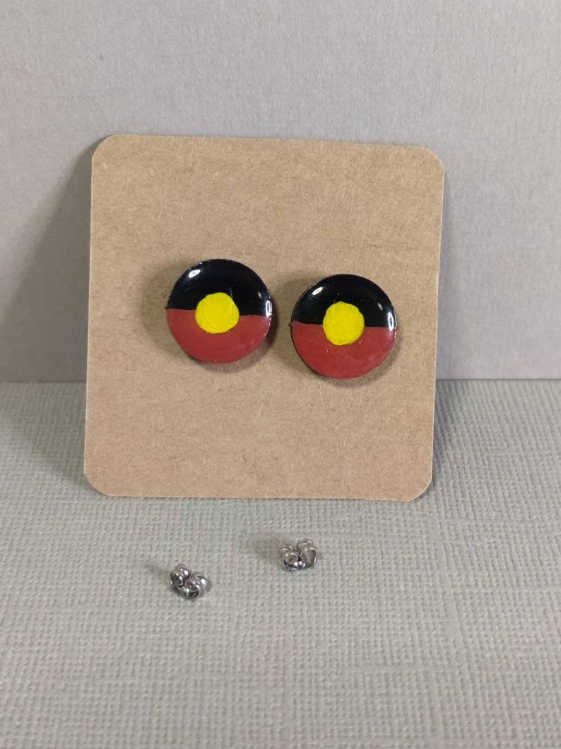 Op de afbeelding: Een paar oorbellen met een zwart, rood en geel Aboriginal vlagontwerp. De oorbellen liggen op een bruine kaart met twee zilveren oorbelsluitingen.