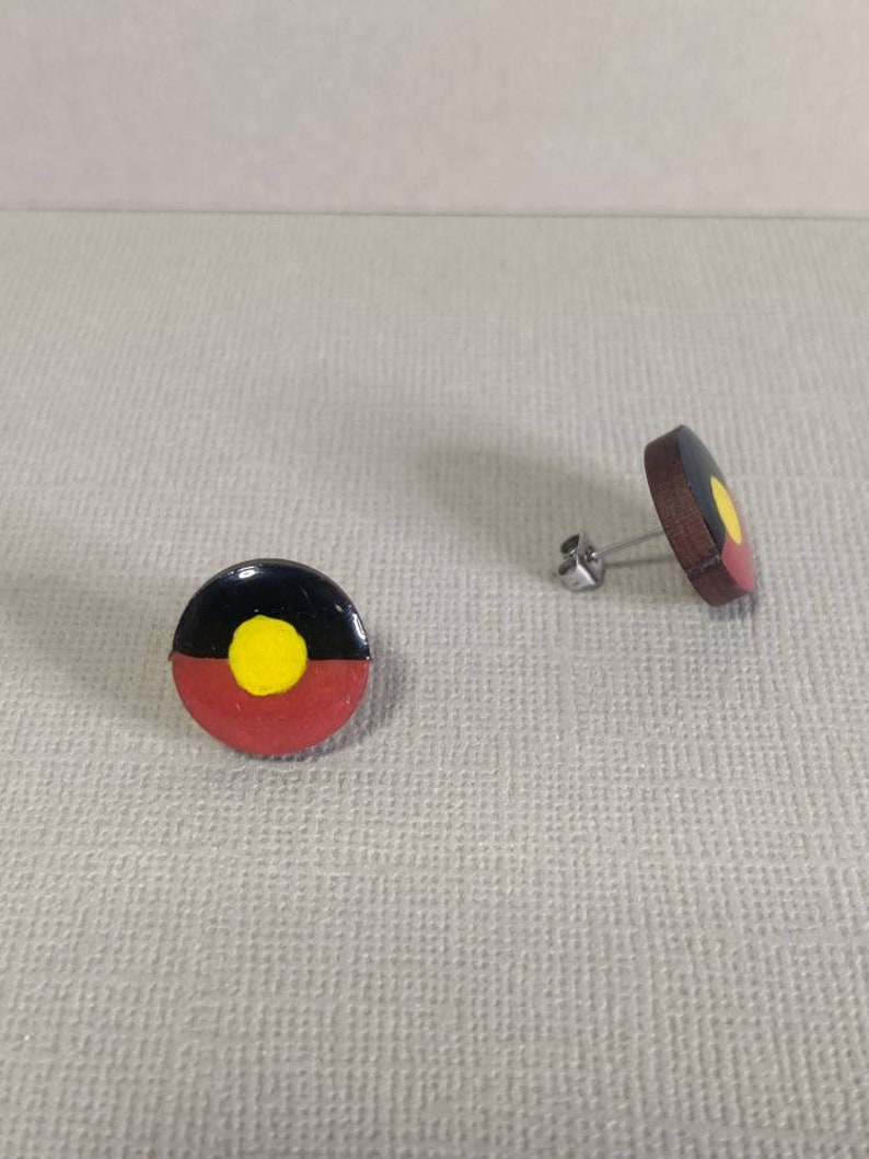 Op de afbeelding: Een paar oorbellen met de Aboriginal vlag. De oorbellen zijn gemaakt van hout en hebben een ronde vorm. De vlag is geschilderd in de traditionele kleuren zwart, rood, geel en wit.