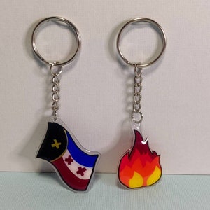 Può includere: Due portachiavi con anelli e catene argentate. Uno presenta un design a bandiera con colori nero, rosso, blu e bianco. L'altro ha un design a fiamma con colori giallo, arancione e rosso.