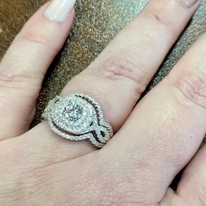 May include: A silver engagement ring with a large, round, center diamond surrounded by smaller diamonds. The ring has a delicate, intricate design with a double halo of diamonds.