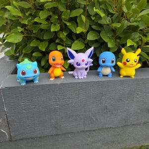 May include: A set of five small plastic character figurines arranged in a row. The collection includes a blue and green creature, an orange bipedal figure, a light purple cat-like character with a red dot, a blue turtle-like figure, and a yellow mouse-like figure.
