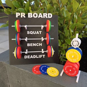 May include: A black PR BOARD with white lettering and colourful weight plates. The board displays "SQUAT", "BENCH", and "DEADLIFT" with barbell designs. Additional weight plates in red, blue, and yellow are also visible.