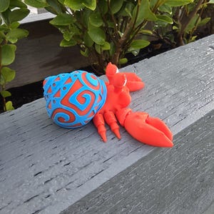 May include: A bright orange and blue crab sculpture. The crab has large orange claws and legs, with a blue ball-shaped body featuring an orange swirl design. The sculpture is on a grey wooden surface, with green foliage in the background.