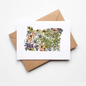 Peut inclure: Une carte de vœux rectangulaire blanche présentant une illustration à l'aquarelle de l'État de l'Oregon remplie de fleurs aux tons jaunes, orange et violets, posée sur une enveloppe en papier kraft marron.