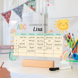 Könnte beinhalten: Ein klarer Wochenplan aus Acryl mit dem Namen "Lisa" und den Wochentagen. Der Plan enthält Fächer wie Mathe, Deutsch und Musik. Auf der rechten Seite befinden sich eine lächelnde Sonne und ein Regenbogen.