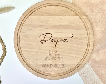 May include: A round wooden cutting board with the text "Papa" and a heart symbol engraved on it. The text below reads "Ein Wort. Zwei Silben. Vier Buchstaben. Unendlich viel Liebe."