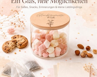 May include: A clear glass jar with a round wooden lid featuring the text "Glücksmomente IM GLAS" and a floral illustration. The jar is filled with pink and white marshmallows and candies, surrounded by cookies, tea bags, nuts, and paper cards.