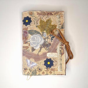 May include: A decorative journal with a tapestry cover featuring floral and leaf appliqués. The cover includes a light blue rose, a green leaf, and two dark blue flower appliqués with yellow centres. A brown leather tie secures the journal.