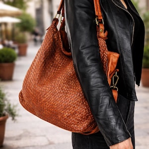 May include: A large, woven leather tote bag in a warm brown hue. The bag features a shoulder strap and a smaller, matching pouch. The bag is carried over the shoulder, paired with a black leather jacket and dark jeans.
