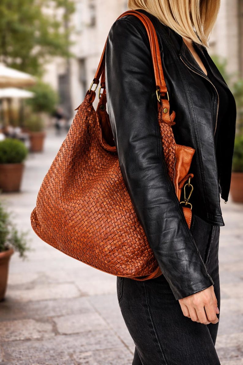 May include: A large, woven leather tote bag in a warm brown hue. The bag features a shoulder strap and a smaller, matching pouch. The bag is carried over the shoulder, paired with a black leather jacket and dark jeans.