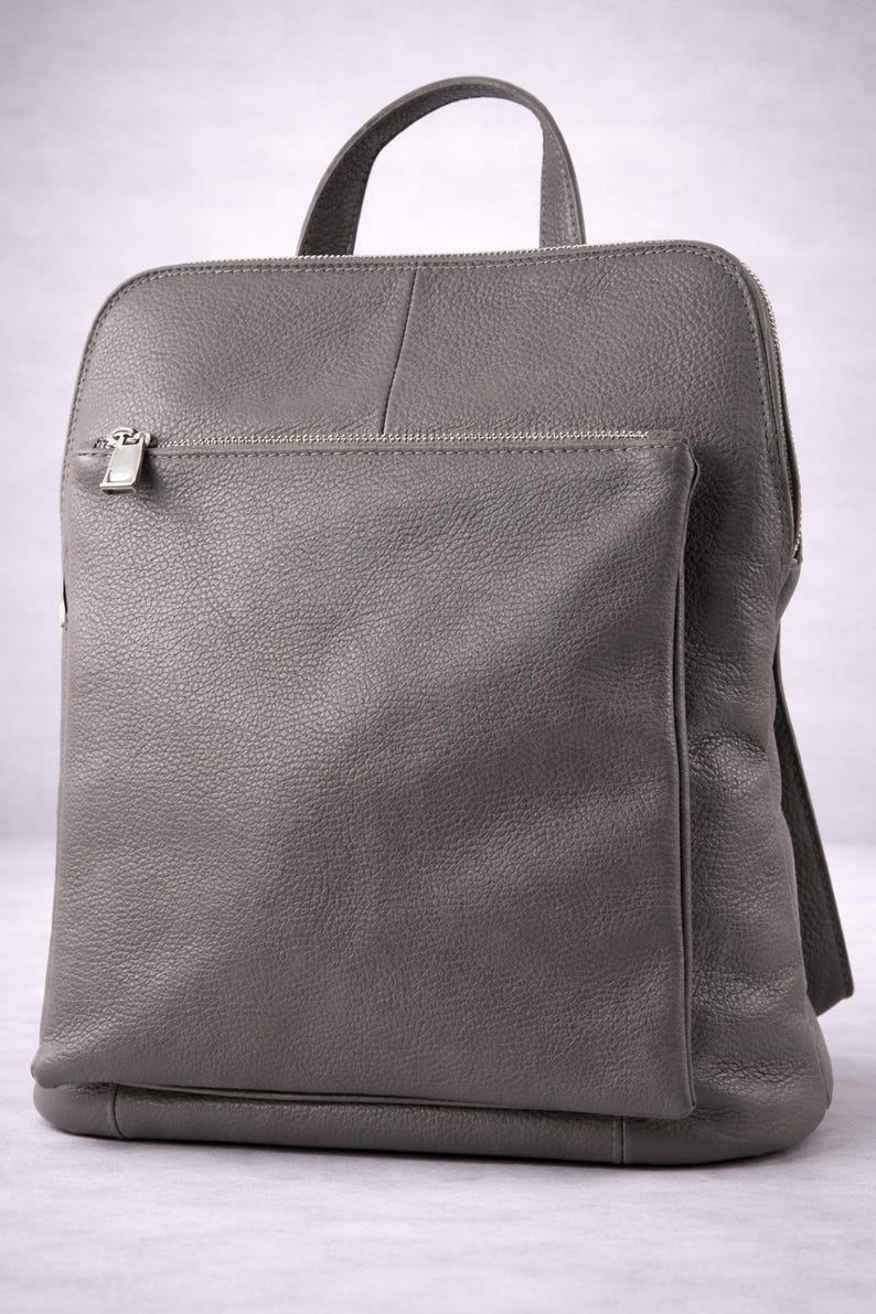K&ouml;nnte beinhalten: Ein grauer Lederrucksack mit einem oberen Griff und einer Rei&szlig;verschlusstasche vorne. Der Rucksack hat eine rechteckige Form mit abgerundeten Ecken und einer strukturierten Oberfl&auml;che. Die Rei&szlig;verschlussanh&auml;nger sind silberfarben.