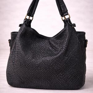 May include: Black woven leather shoulder bag with two handles and gold-colored hardware. The bag has a large, open top and a woven texture throughout the body. The handles are attached with gold-colored rings.