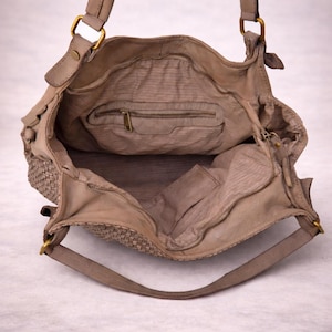 May include: Open view of a beige leather handbag with a woven side panel. The bag has a top handle and a long shoulder strap. The interior features a zippered pocket and a small open pocket. Gold-colored hardware accents the bag.
