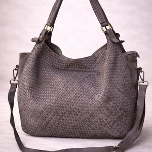 May include: A gray woven leather handbag with a shoulder strap and top handles. The bag features a woven pattern on the main body and is accented with brass-colored hardware. The bag has a large capacity and a casual style.