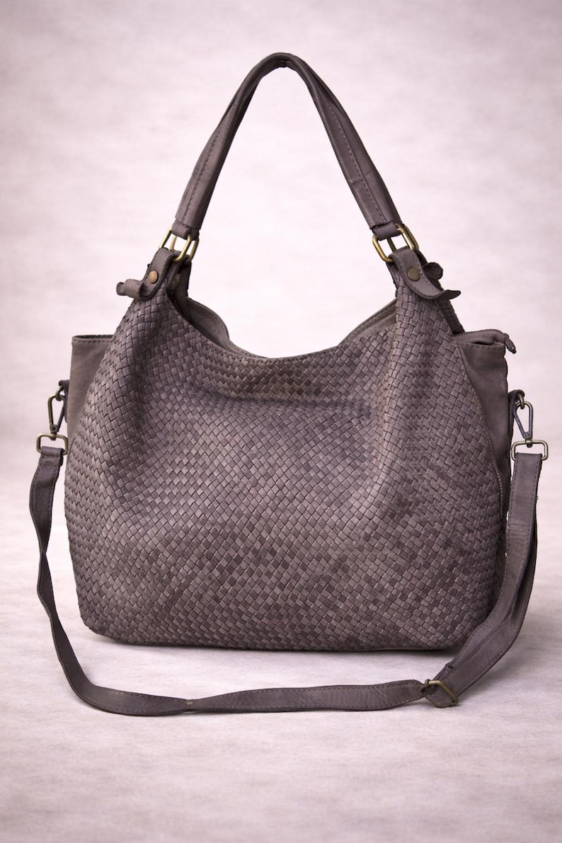 May include: A gray woven leather handbag with a shoulder strap and top handles. The bag features a woven pattern on the main body and is accented with brass-colored hardware. The bag has a large capacity and a casual style.