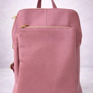 K&ouml;nnte beinhalten: Ein staubrosa Rucksack mit einem oberen Griff und einer Vordertasche. Die Tasche hat goldfarbene Rei&szlig;verschl&uuml;sse und Beschl&auml;ge. Das Material scheint ein strukturiertes Leder oder leder&auml;hnliches Material zu sein.