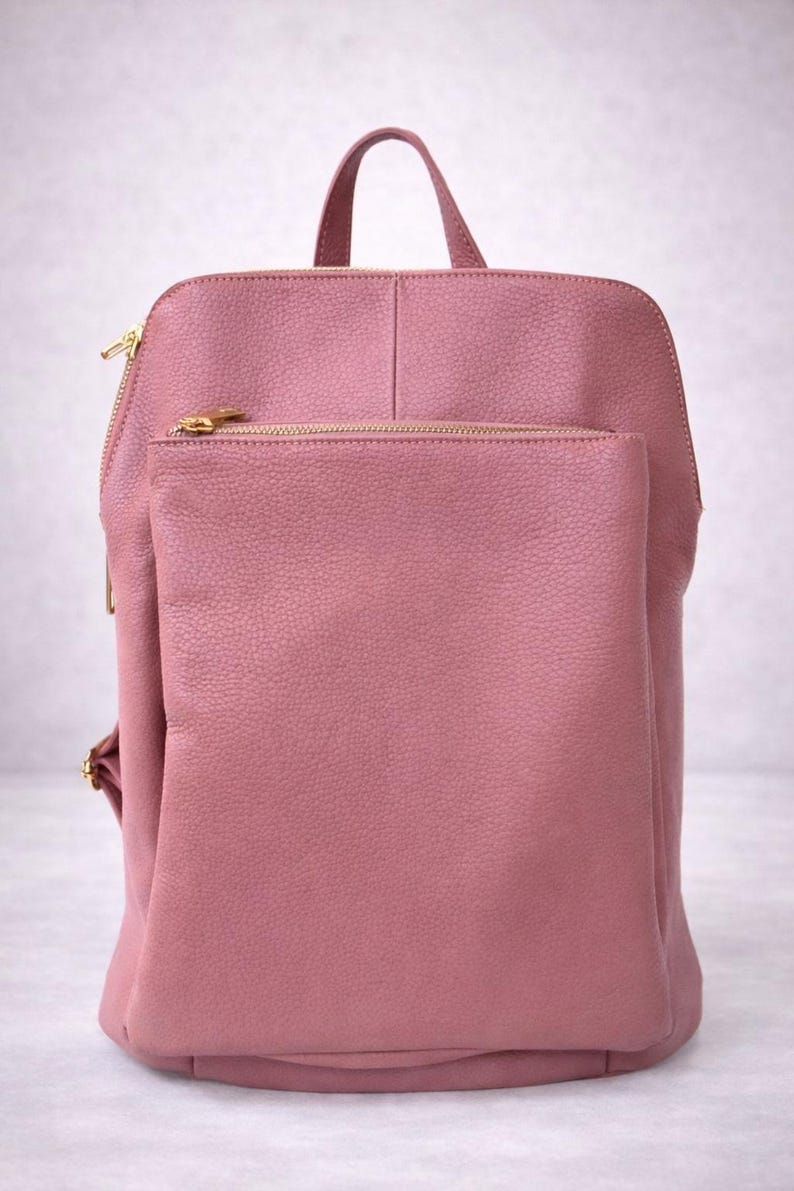 K&ouml;nnte beinhalten: Ein staubrosa Rucksack mit einem oberen Griff und einer Vordertasche. Die Tasche hat goldfarbene Rei&szlig;verschl&uuml;sse und Beschl&auml;ge. Das Material scheint ein strukturiertes Leder oder leder&auml;hnliches Material zu sein.
